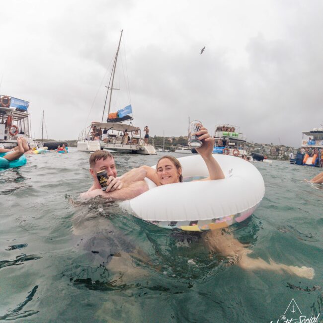 Two people relax in the ocean, smiling and holding drinks. One lounges in a white inflatable ring, while the other leans on it. Boats and other people are visible in the background under a cloudy sky.