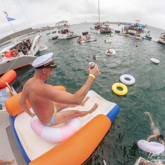 A man in a captain’s hat sits on an inflatable float with a drink, about to slide into the water at a lively boat party. People swim and relax on floats, with several boats anchored nearby.