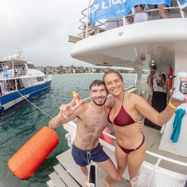 A smiling man and woman in swimwear stand on a boat deck, posing for a photo. The man holds up a peace sign. Other people and another boat are visible in the background on the water.