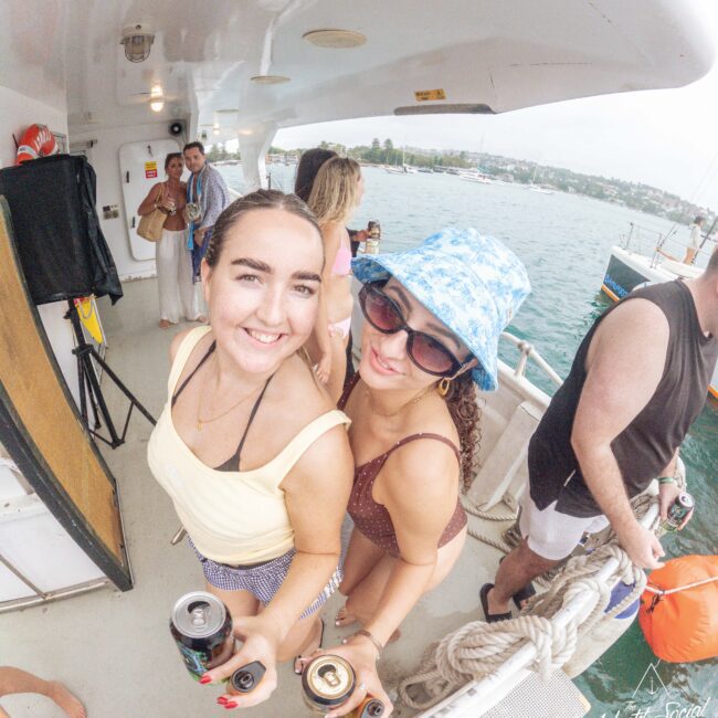 Two women smiling at the camera while holding drinks on a boat. One wears a yellow top and the other wears a blue bucket hat and sunglasses. The boat is on water, with other people and land visible in the background.