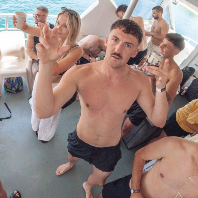 A shirtless man in black swim trunks poses with his hands up, holding a drink, among other people on a boat. The group appears to be enjoying a sunny day on the water.