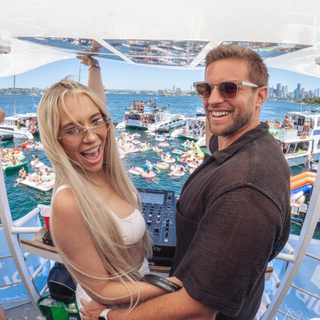 A man and woman smile at the camera while standing at a DJ booth on a boat. Behind them, numerous boats and people are gathered on the water, enjoying a lively summer party. The city skyline is visible in the background.