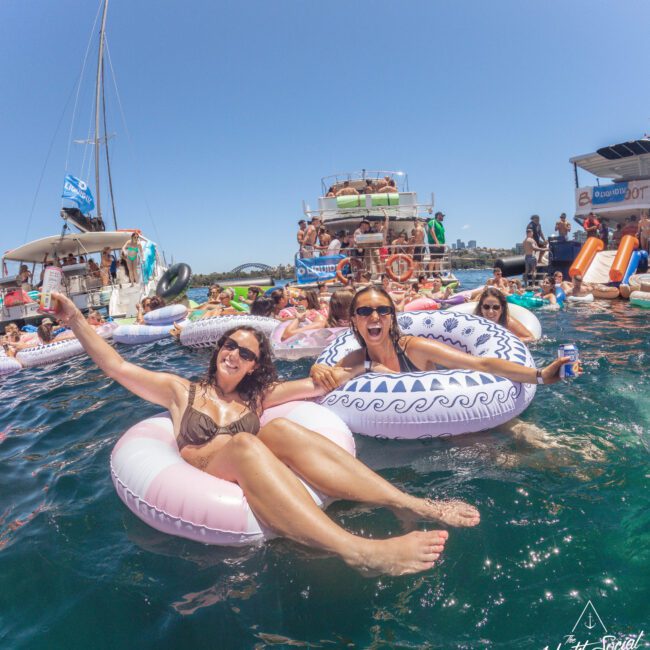 Two women in sunglasses smile and hold drinks while floating on inflatable pool rings in the water, surrounded by other people and boats on a sunny day. A festive, lively summer scene.