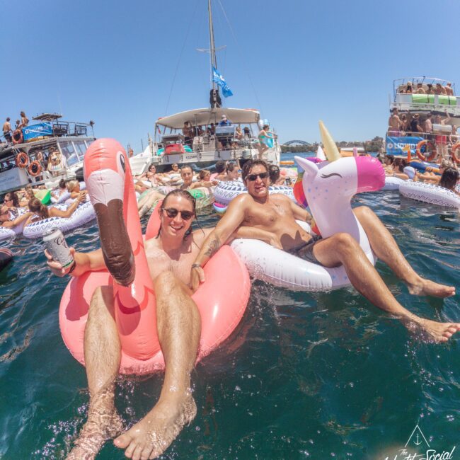 Two men relax on inflatable floats shaped like a flamingo and a unicorn in the ocean, surrounded by other people on floats and boats, enjoying a sunny day at a lively yacht party.
