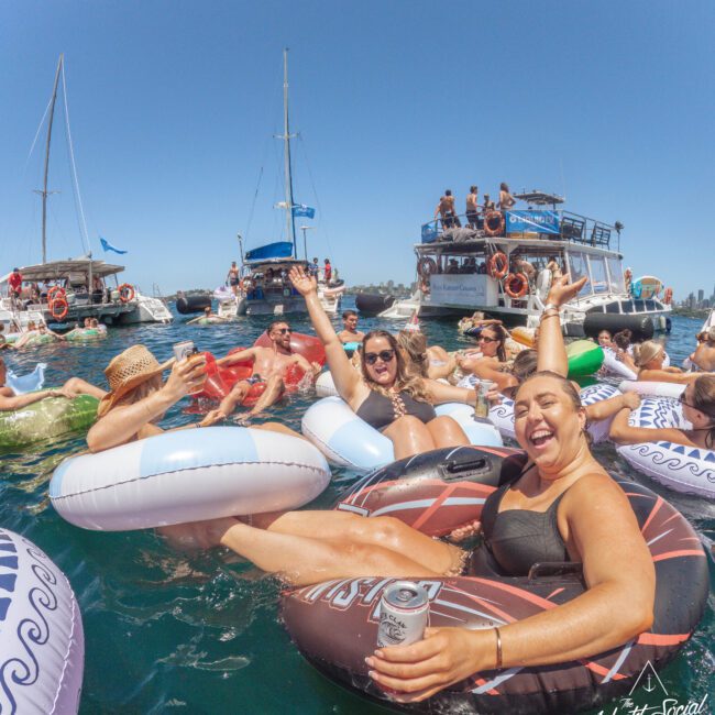 A group of people relax on colorful pool floats in the water, smiling and raising drinks. Boats and more people are in the background under a clear blue sky, creating a lively party atmosphere.