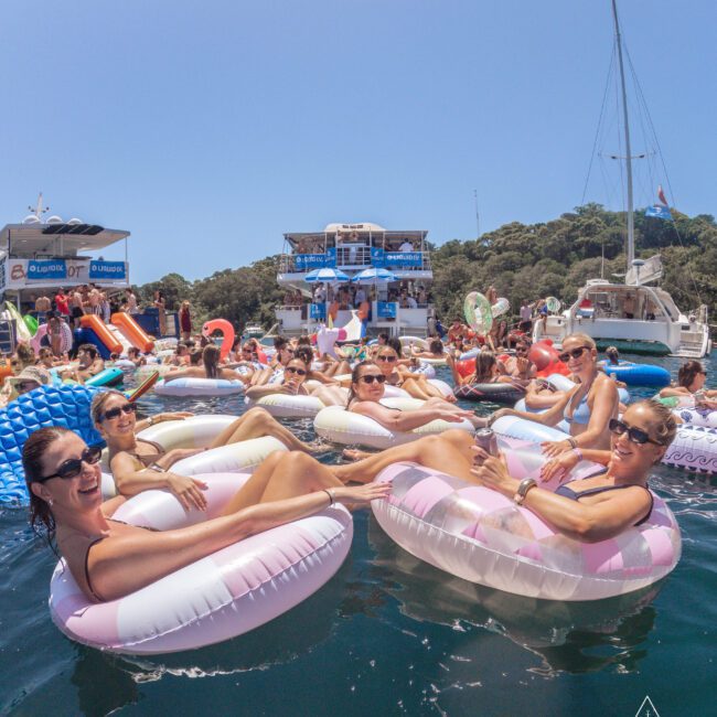 A group of people relaxing on inflatable rings in the water, smiling and enjoying a sunny day near boats and yachts, with blue skies and tree-covered hills in the background.