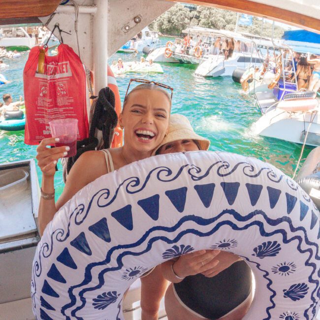 Two women on a boat smile at the camera, one holding a drink and a large blue-and-white inflatable ring. People swim and relax on boats in the turquoise water in the background.