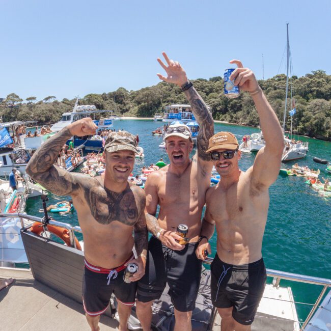 Three shirtless men with tattoos and sunglasses pose smiling on a boat, holding drinks. Behind them, people float on the water and several boats are anchored near a forested shoreline under a sunny sky.