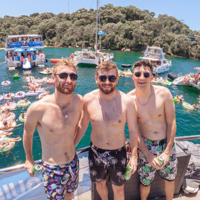 Three men in swim trunks stand smiling on a dock holding drinks, with people swimming and lounging on floats in the water behind them. Boats and tree-covered land are visible in the background on a sunny day.