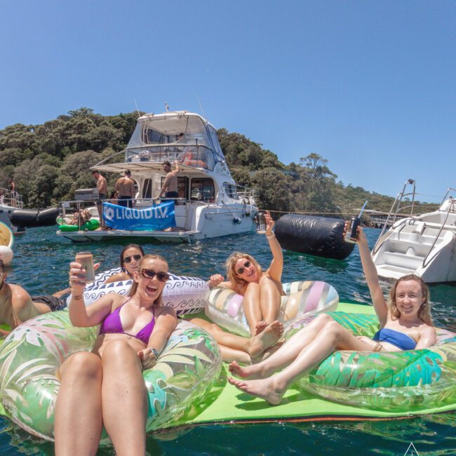 Four women in swimsuits relax on inflatable floats in the water, smiling and raising drinks. A yacht and trees are in the background under a sunny sky. The atmosphere is festive and summery.