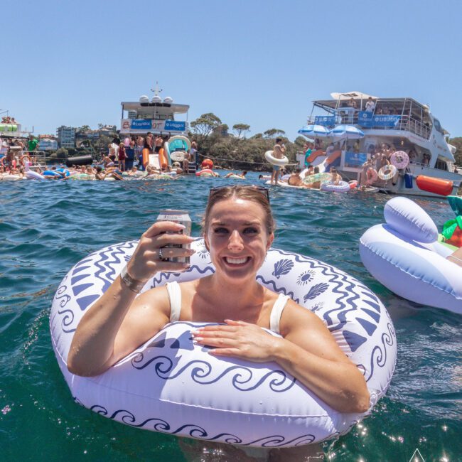 A smiling woman in a patterned inflatable ring holds up a drink while floating in the water. There are people on boats and in the water, with inflatables and a sunny sky in the background.