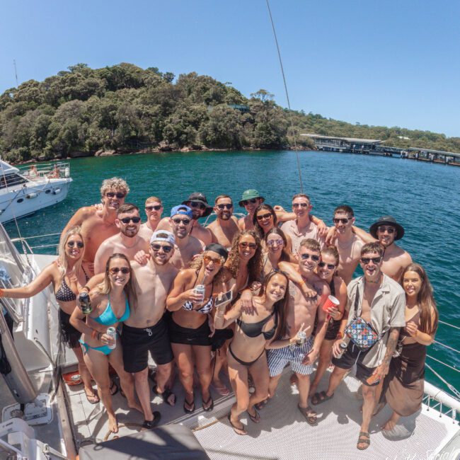 A group of young adults in swimwear pose and smile together on a boat with clear blue water and a green, tree-covered island in the background under a sunny sky.