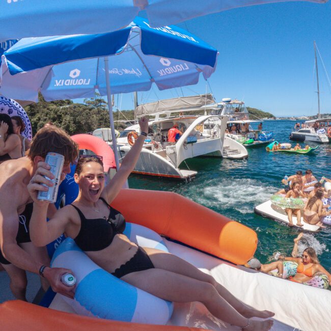 A woman in a black bikini lounges on a pool float, smiling and raising a drink, surrounded by people on boats and inflatables at a lively lake party under a blue sky and umbrellas.
