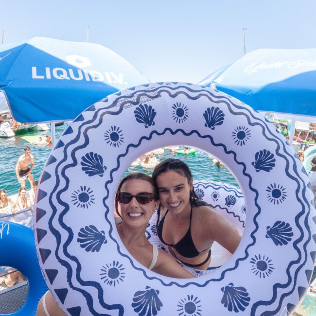 Two women in swimsuits smile through a large blue and white pool float, with a group of people swimming and relaxing on a boat in the background under blue "Liquid IV" umbrellas on a sunny day.