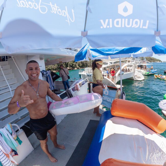 A smiling man in swim trunks holds a pink and white pool float on a boat deck, with people relaxing and swimming nearby. Other boats and green trees are visible in the sunny background.