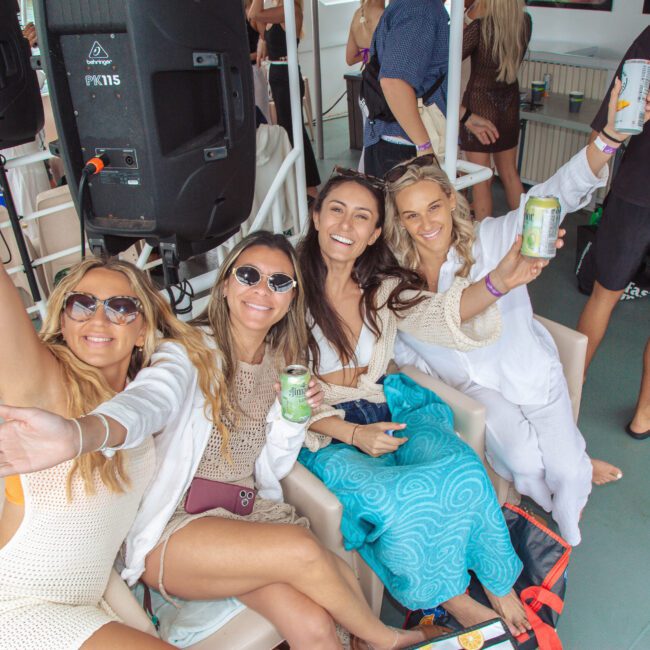 Four women sit together smiling and holding drinks on a boat, dressed in summer outfits and sunglasses. One woman waves at the camera; others pose closely. The background shows other people and speakers, capturing a lively atmosphere.
