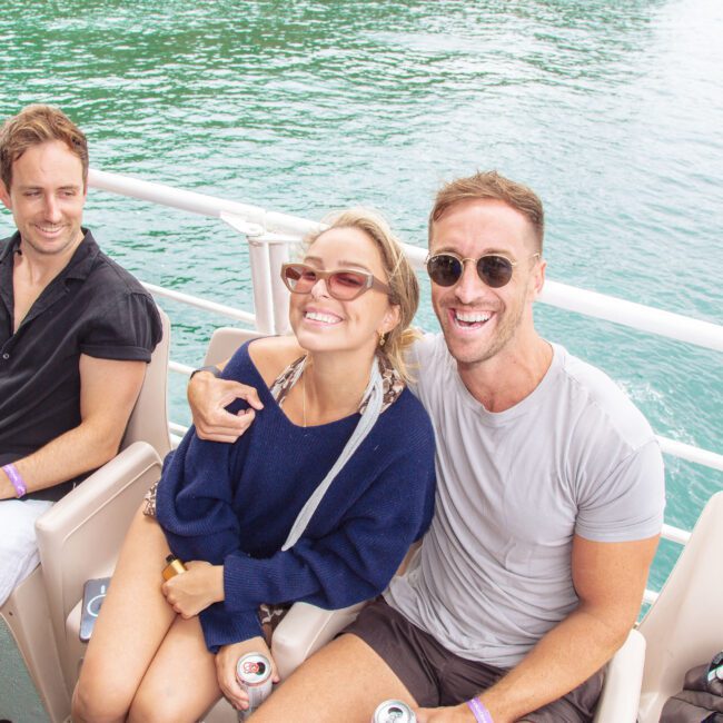 Three people sit on a boat by turquoise water, smiling and enjoying drinks. The two on the right, a woman in sunglasses and a man in a white shirt, pose closely, while the third person sits to their left, also smiling.