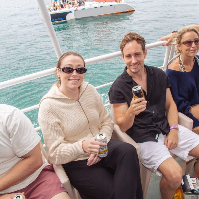 Four people sit on a boat, smiling and holding canned drinks. The water and another boat are in the background. They appear relaxed, wearing casual summer clothing and sunglasses.