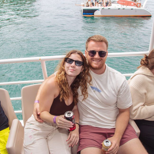 A smiling man and woman sit close together on a boat, both wearing sunglasses and holding drinks. Other people are seated nearby, and turquoise water with another boat is visible in the background.