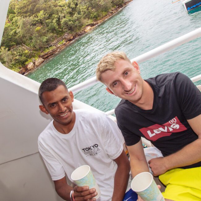 Two men smiling and holding cups sit on a boat with water and green trees in the background. The man on the left wears a white shirt; the man on the right wears a black Levi’s shirt and yellow shorts.