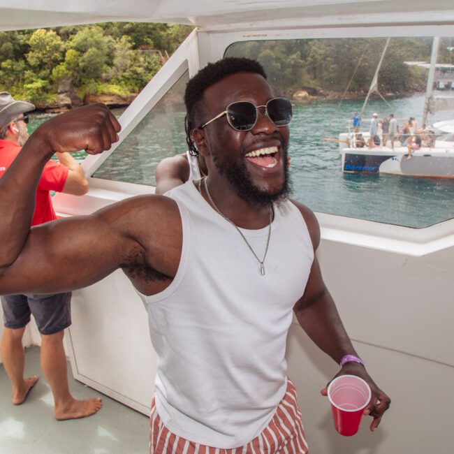 A smiling man in sunglasses and a white tank top flexes his arm on a boat, holding a red cup. Other people are on the boat and another boat with people is visible in the background on the water.