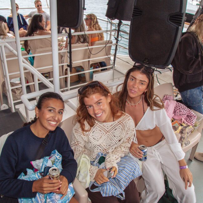 Three women smile at the camera while sitting on a boat, holding drinks. Other people are mingling in the background, and the logo "Yacht Social Club" is visible in the corner of the image.