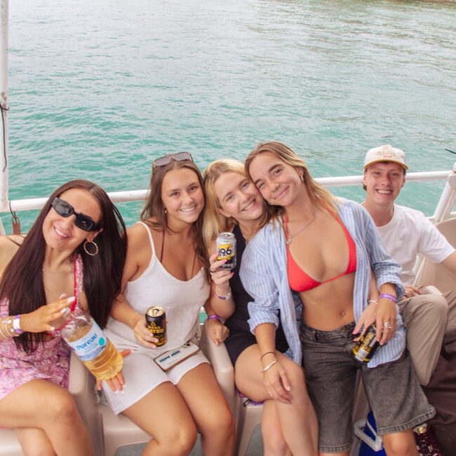 Five young adults sit smiling on a boat, holding drinks and enjoying a sunny day on the water. The ocean and shoreline are visible in the background. Everyone appears relaxed and happy.