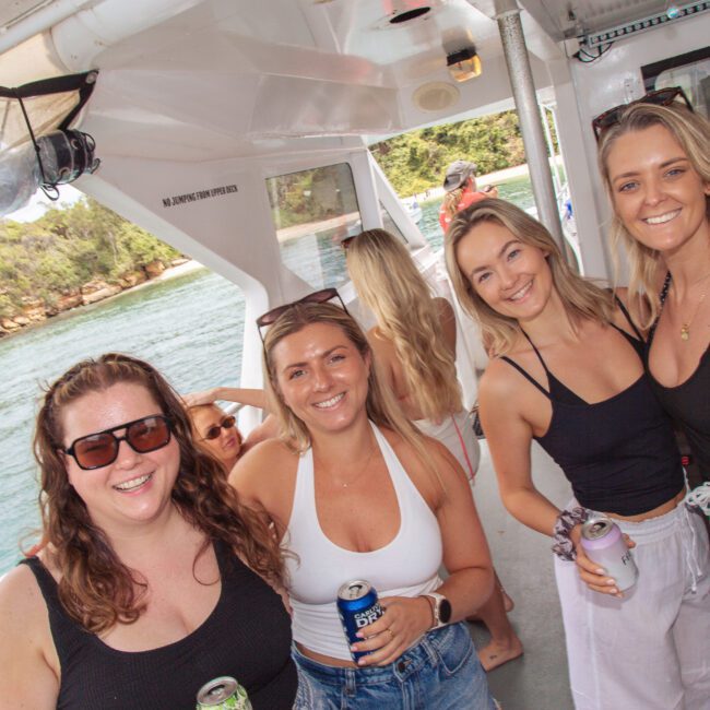 Four women smiling and holding drinks on a boat, with water and trees visible outside. They are casually dressed and enjoying a sunny day, with more people socializing in the background.