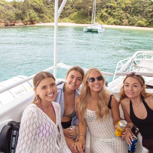 Four women smiling and sitting on a boat with drinks in hand. Behind them is clear blue-green water, a docked catamaran, and a lush, tree-lined shore under a cloudy sky.