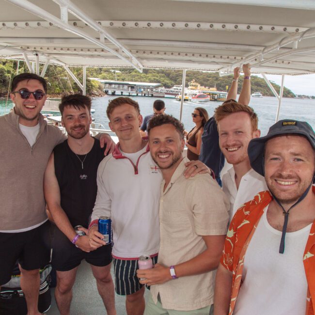 Six smiling men stand together on a boat, posing for a photo. The background shows water, trees, and buildings under a partly cloudy sky. Some hold drinks, and one wears a bucket hat and orange patterned shirt.