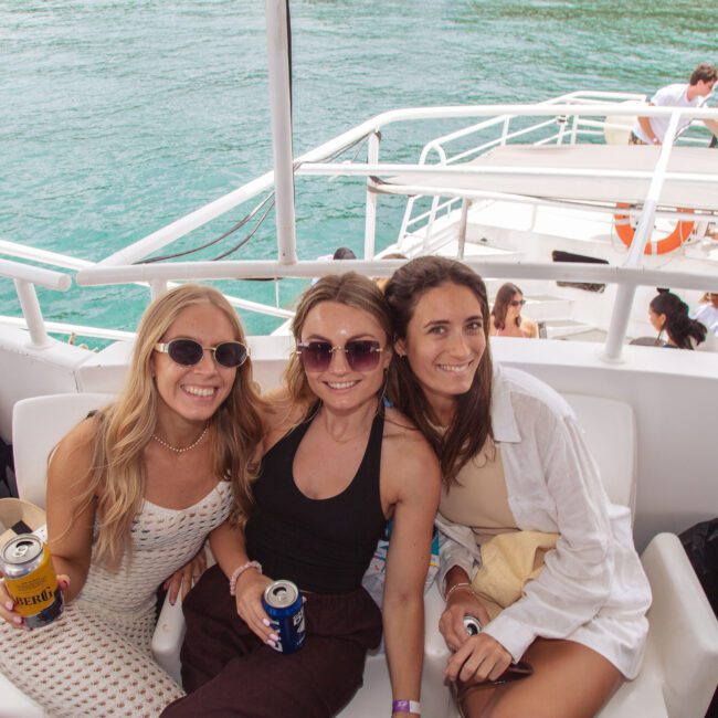 Three women smiling and sitting together on a boat, each holding a drink, with clear turquoise water and other people socializing on the lower deck in the background.