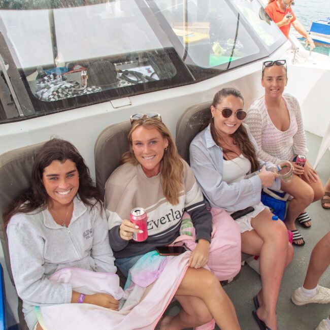 Four women sit and smile on a boat, holding drinks and wearing casual clothes. The sea and other people are visible in the background, indicating a relaxed, social atmosphere.