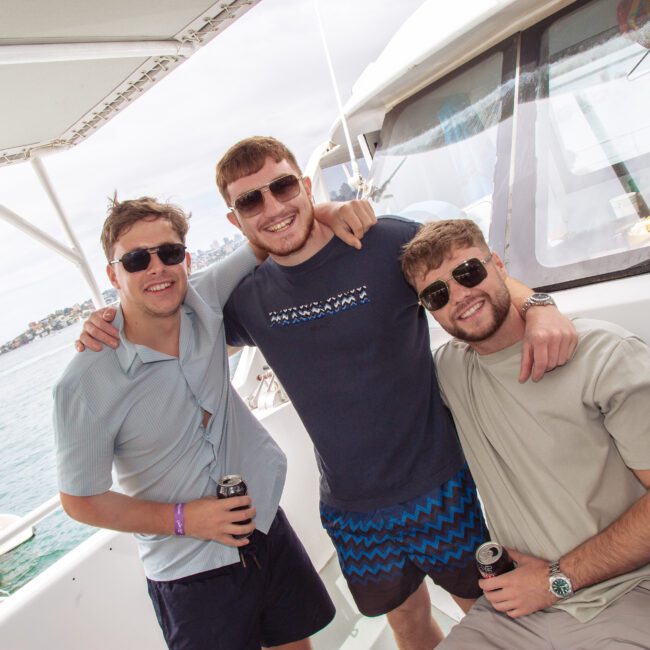 Three young men wearing sunglasses and casual outfits smile and pose together on a boat, each holding a drink. In the background, water, land, and other people on boats are visible under a cloudy sky.