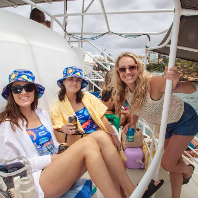 Three women on a boat, two sitting and wearing matching blue bucket hats and swimsuits, one standing and smiling at the camera. They are relaxing, holding drinks, with water and a rocky shoreline in the background.