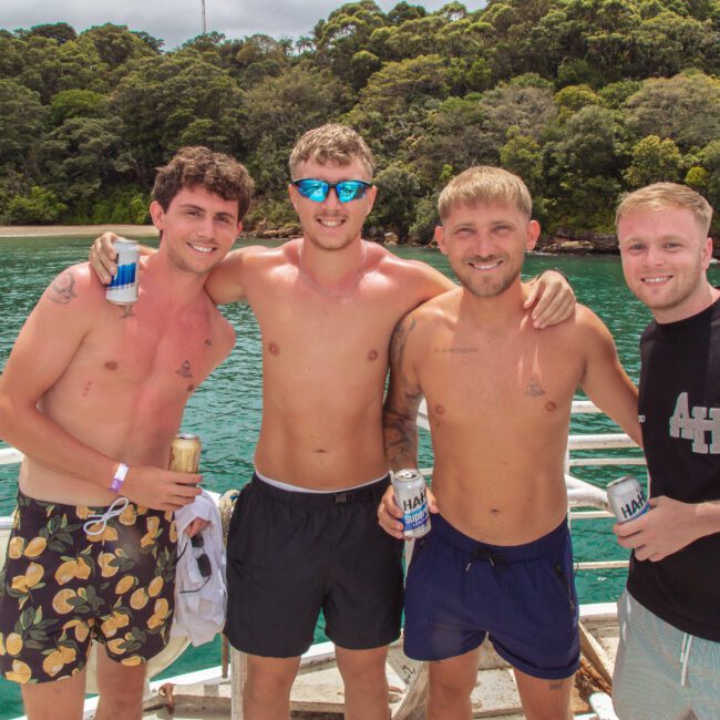 Four young men in swimwear stand arm-in-arm on a boat, smiling at the camera. They hold cans of drinks, with green water and lush trees in the background on a sunny day.
