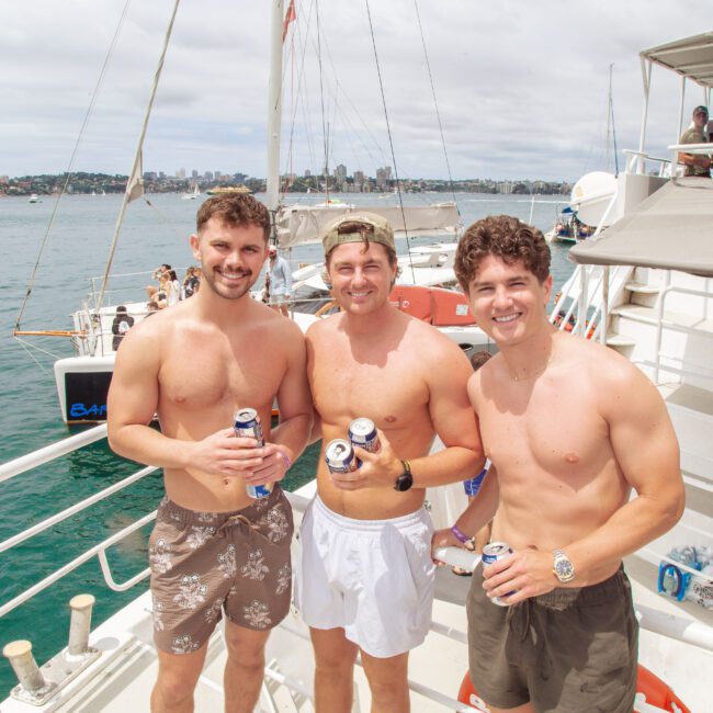 Three young men in swim trunks stand on a boat deck holding canned drinks, smiling at the camera. The ocean and city skyline are visible in the background under a partly cloudy sky.