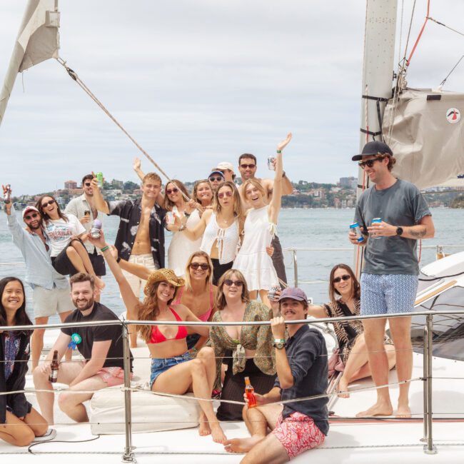 A group of people pose and smile on the deck of a sailboat, some holding drinks. The boat is on the water with a coastal town visible in the background under a cloudy sky.