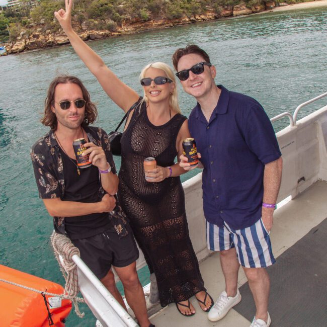 Three people stand on a boat, smiling and holding drinks. One woman, wearing sunglasses and a black outfit, raises her arm cheerfully. Two men stand beside her, both wearing sunglasses and striped shorts. Water and boats are visible in the background.
