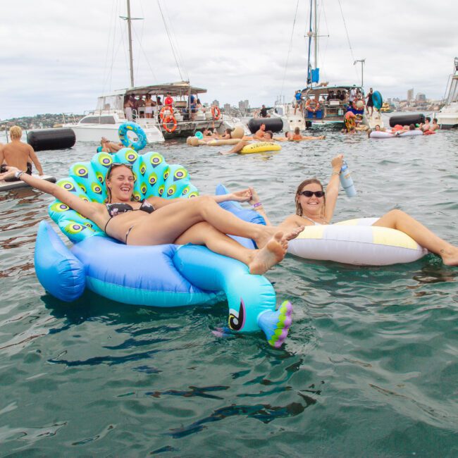 Two women relaxing on inflatable floats in the water, surrounded by other people on floats and boats. The woman on the left is on a blue peacock float, both are smiling and enjoying a social event on the water.