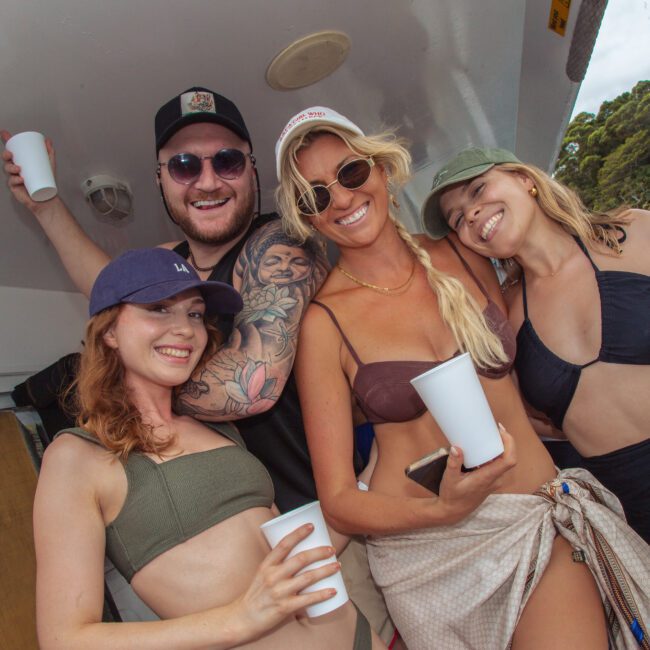Four smiling people in swimsuits pose together on a boat, each holding a white cup. Trees and water are visible in the background, suggesting a fun outdoor gathering or party.