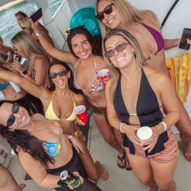 A group of women in swimsuits and sunglasses smile, laugh, and pose for a photo on a boat. Some are holding drinks, and there is water visible in the background. The mood is festive and celebratory.