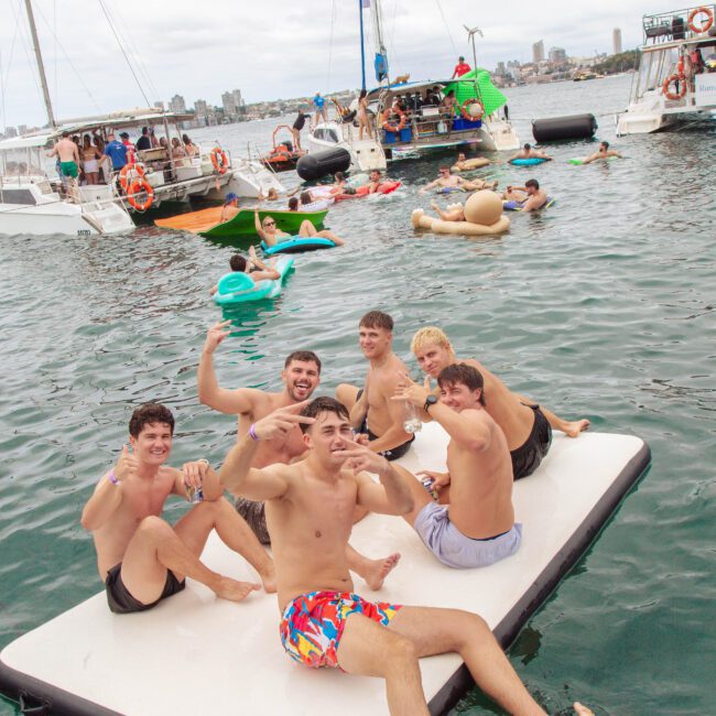 A group of young men in swimwear smile and gesture at the camera while sitting on a floating platform in the water, with boats and other people swimming and relaxing in the background.