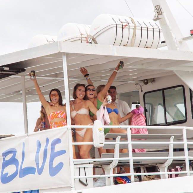 Four young adults in swimwear smile and celebrate, holding drinks on the upper deck of a white boat. A large banner reading "BLUE" hangs from the railing. A crew member in red stands nearby under a cloudy sky.