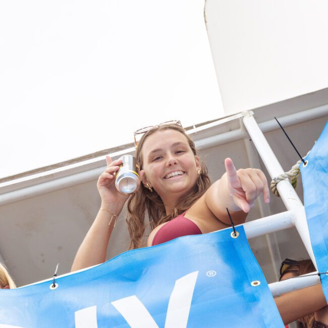 A young woman in a bikini top smiles and points at the camera while holding a drink can, standing behind a blue banner on a boat. Another person is partially visible beside her.