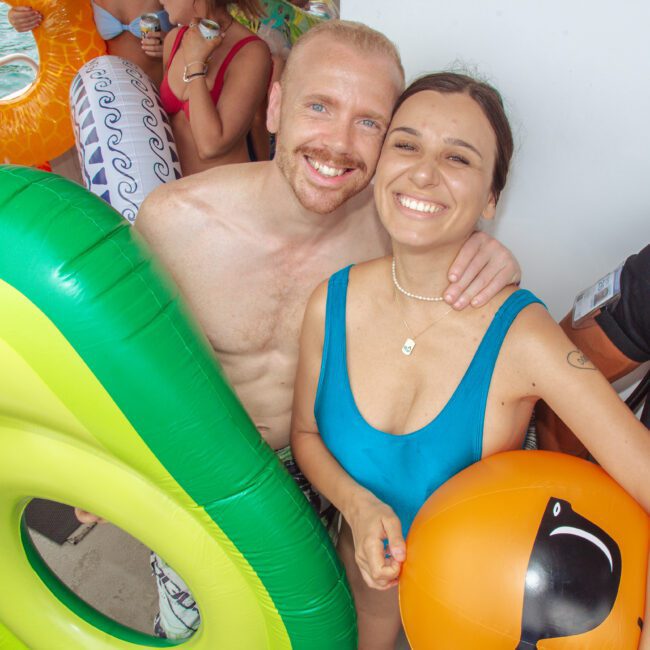 Two people in swimsuits smile at the camera on a boat, holding inflatable pool floats including a green avocado and orange beach ball. Other people and floats are visible in the background, with water nearby.