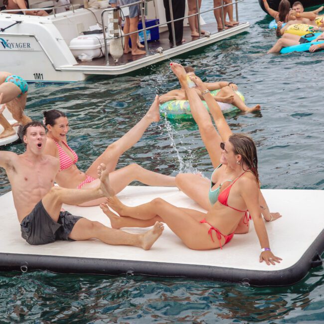 Four young adults in swimsuits sit on a floating mat in the water, playfully splashing each other; boats and more people on inflatables are visible in the background, creating a lively summer scene.