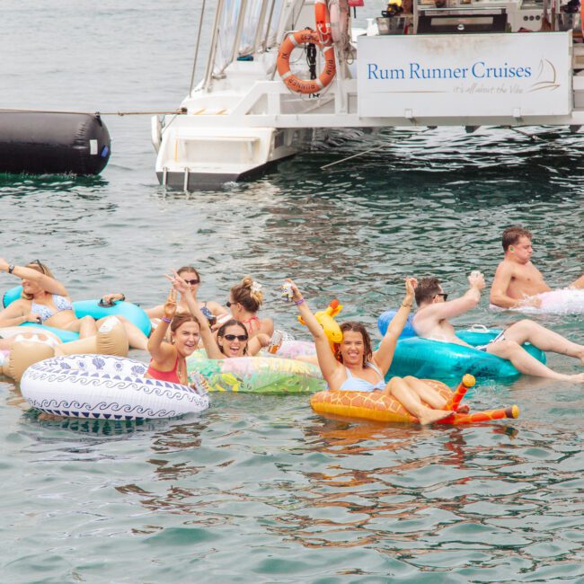 A group of people in swimsuits relax on colorful inflatable floats near a boat labeled "Rum Runner Cruises," enjoying a sunny day on the water. Some smile and wave at the camera.