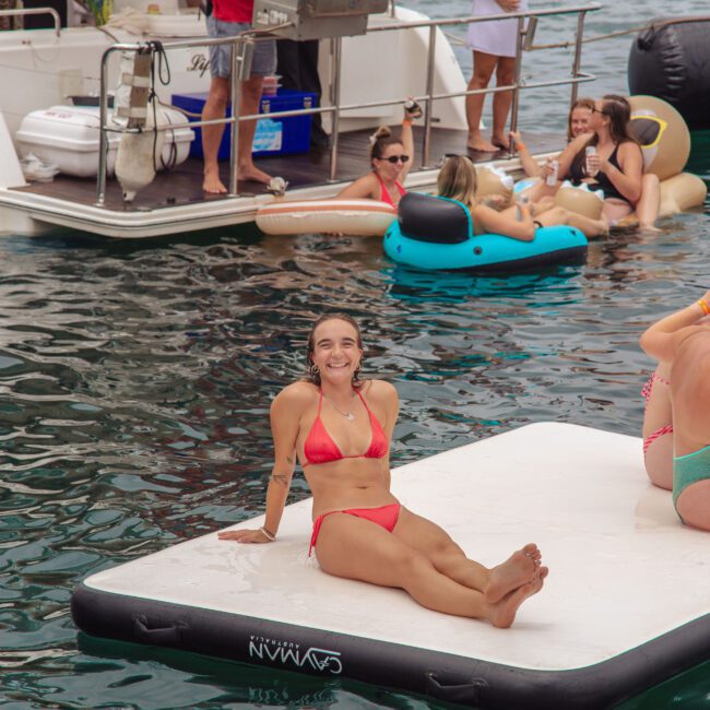 A woman in a red bikini smiles while sitting on a floating mat in the water, surrounded by others on inflatables and a nearby boat. People are socializing and grilling on the boat in the background.