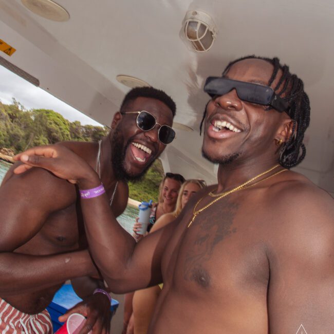 Two men wearing sunglasses and swim trunks laugh together on a boat, surrounded by friends holding drinks. Trees and water are visible in the background. The atmosphere is lively and joyful.