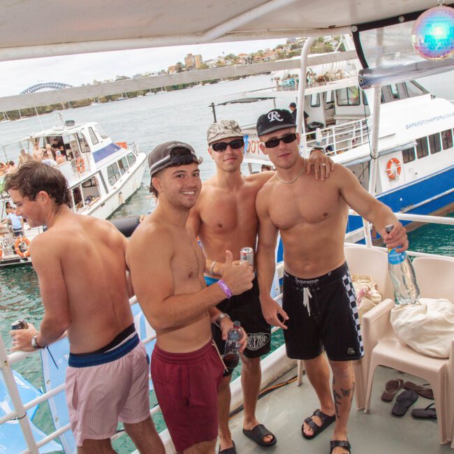 Four young men in swimwear stand on a boat, smiling at the camera. Other boats and people are visible in the background, and the scene appears festive and relaxed on the water.
